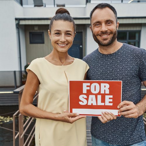 Happy man and woman standing with a sign for sale in front of a big house and looking friendly